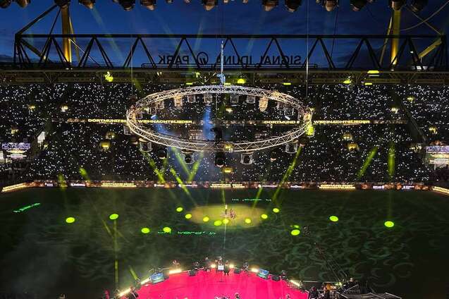 Luftaufnahme eines Stadions bei Nacht mit leuchtenden Lichtern und einer zentralen B&uuml;hnenkonstruktion auf dem Spielfeld.