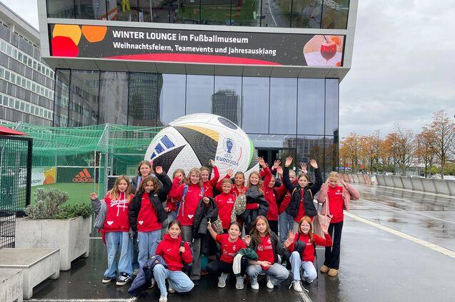 Gruppe von M&auml;dchen in roten T-Shirts posiert vor einem gro&szlig;en Fu&szlig;ball und dem Fu&szlig;ballmuseum, l&auml;chelnd und winkend.
