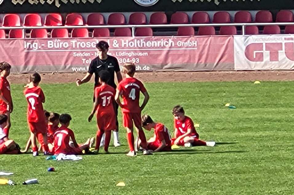 Gruppe von Kindern in roten Trikots sitzt auf dem Fu&szlig;ballplatz, Trainer erkl&auml;rt &Uuml;bungen, Zuschauer im Hintergrund.