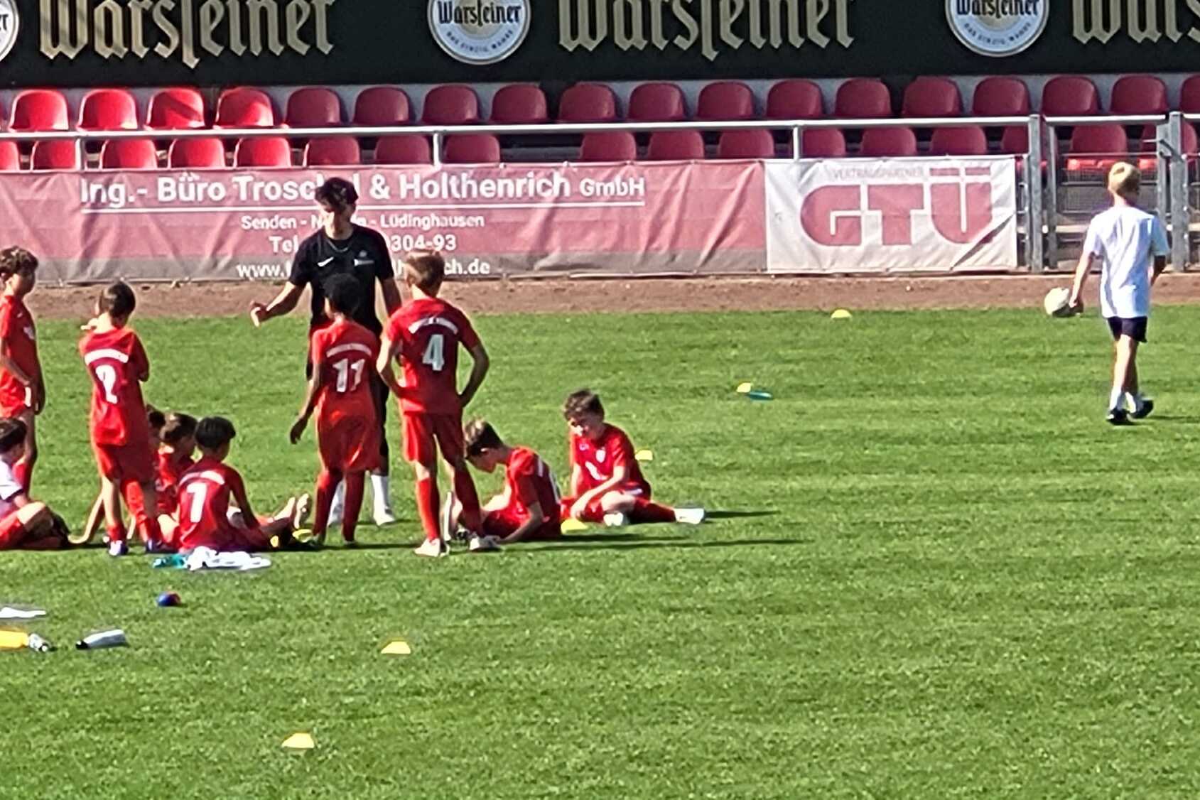 Gruppe von Kindern in roten Trikots sitzt auf dem Fu&szlig;ballplatz, Trainer erkl&auml;rt &Uuml;bungen, Zuschauer im Hintergrund.