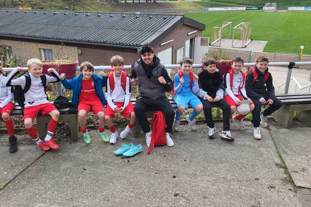 Gruppe von zehn jungen Fu&szlig;ballspielern, sitzend auf einer Bank, im Hintergrund ein Fu&szlig;ballfeld und ein Sportgeb&auml;ude.