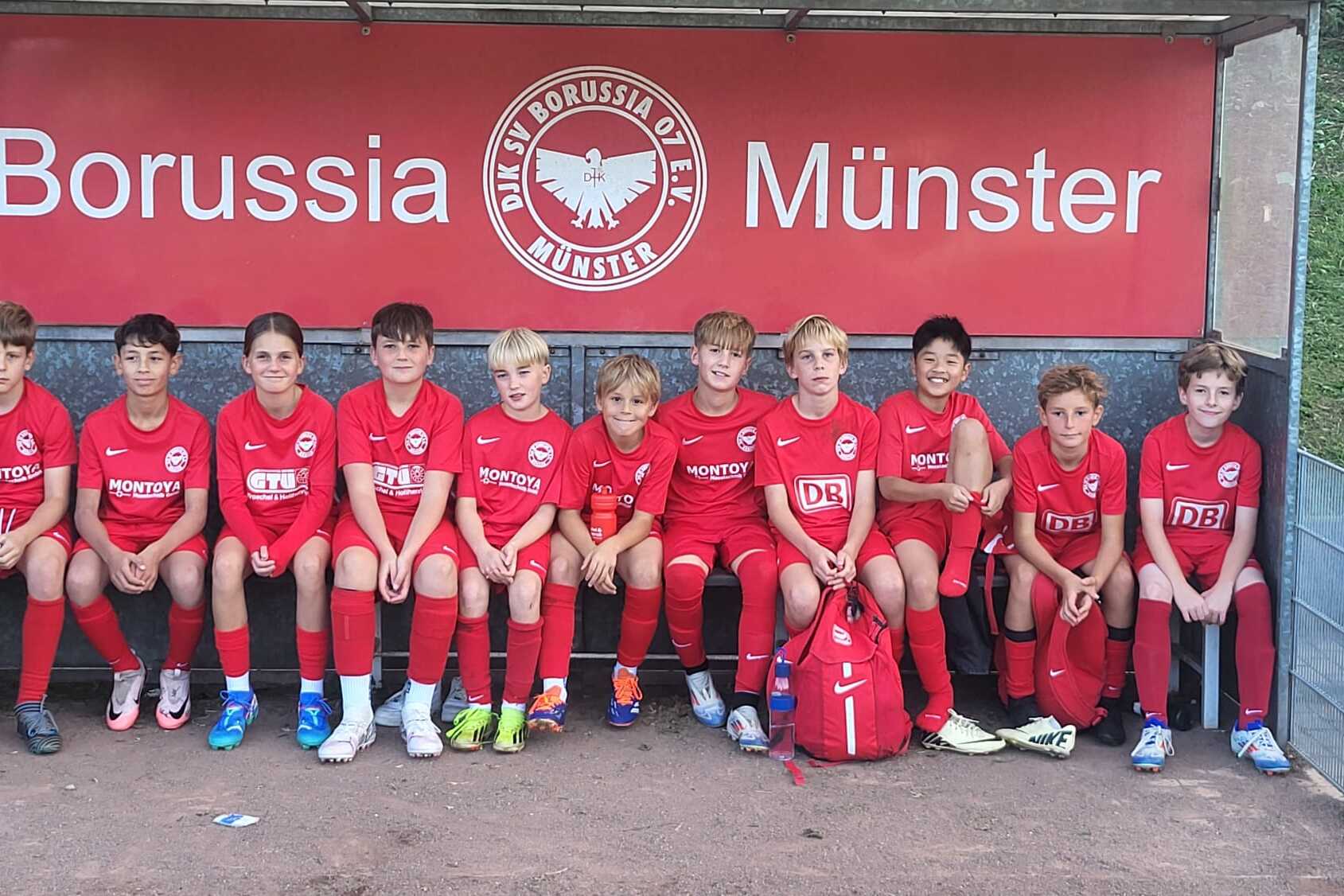 Zehn Kinder in roten Fu&szlig;balltrikots sitzen auf einer Bank unter einem Borussia M&uuml;nster-Schild.