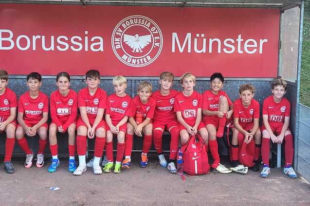Zehn Kinder in roten Fu&szlig;balltrikots sitzen auf einer Bank unter einem Borussia M&uuml;nster-Schild.