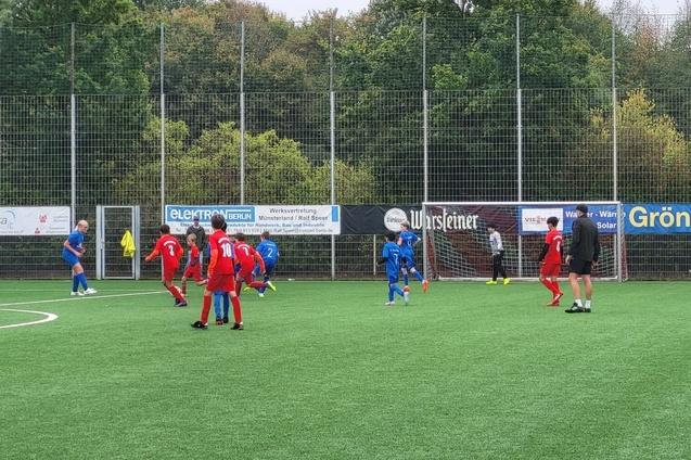 Jugend-Fu&szlig;ballspiel auf einem Kunstrasenplatz mit Spielern in roten und blauen Trikots, Torwart im Hintergrund.