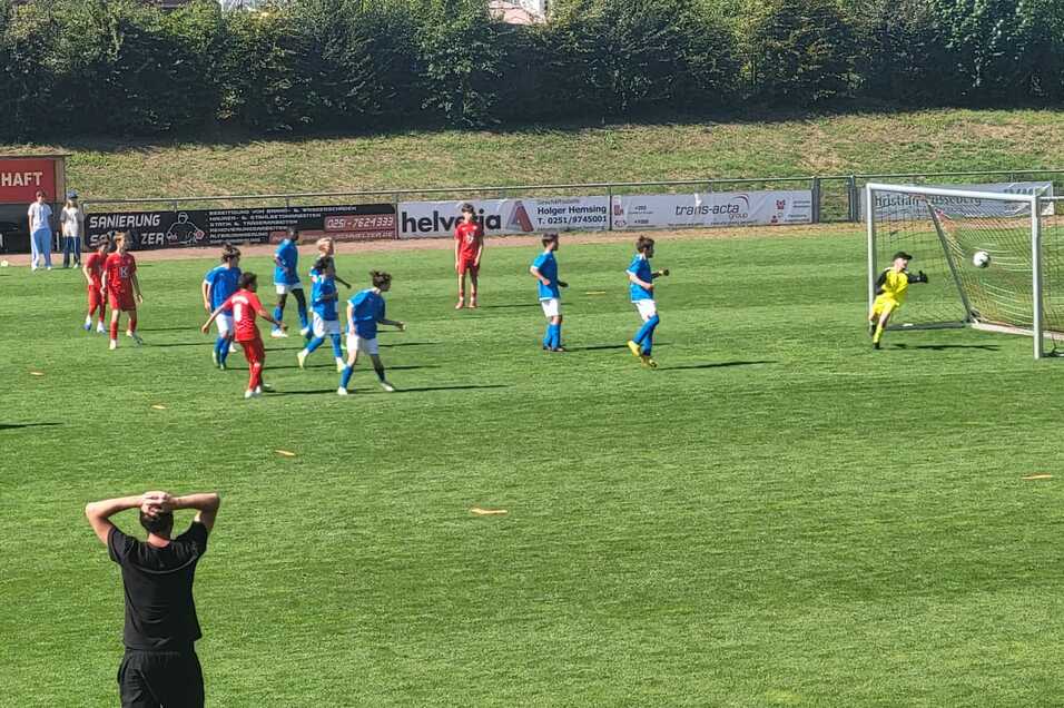 Spielszene mit Kindern in blauen und roten Trikots beim Elfmeter, Zuschauer beobachten gespannt vom Spielfeldrand.
