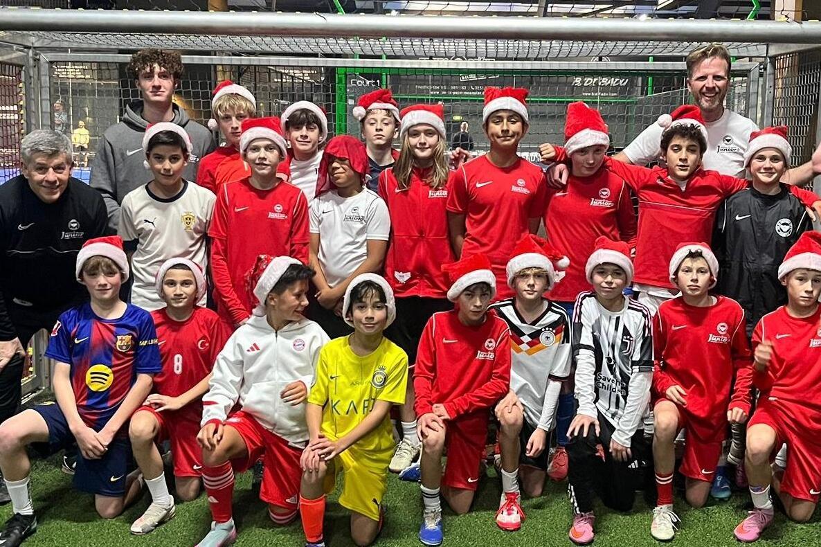 Gruppierung von 20 Kindern in Fußballtrikots und Weihnachtsmützen vor einem Tor auf einem Sportplatz.