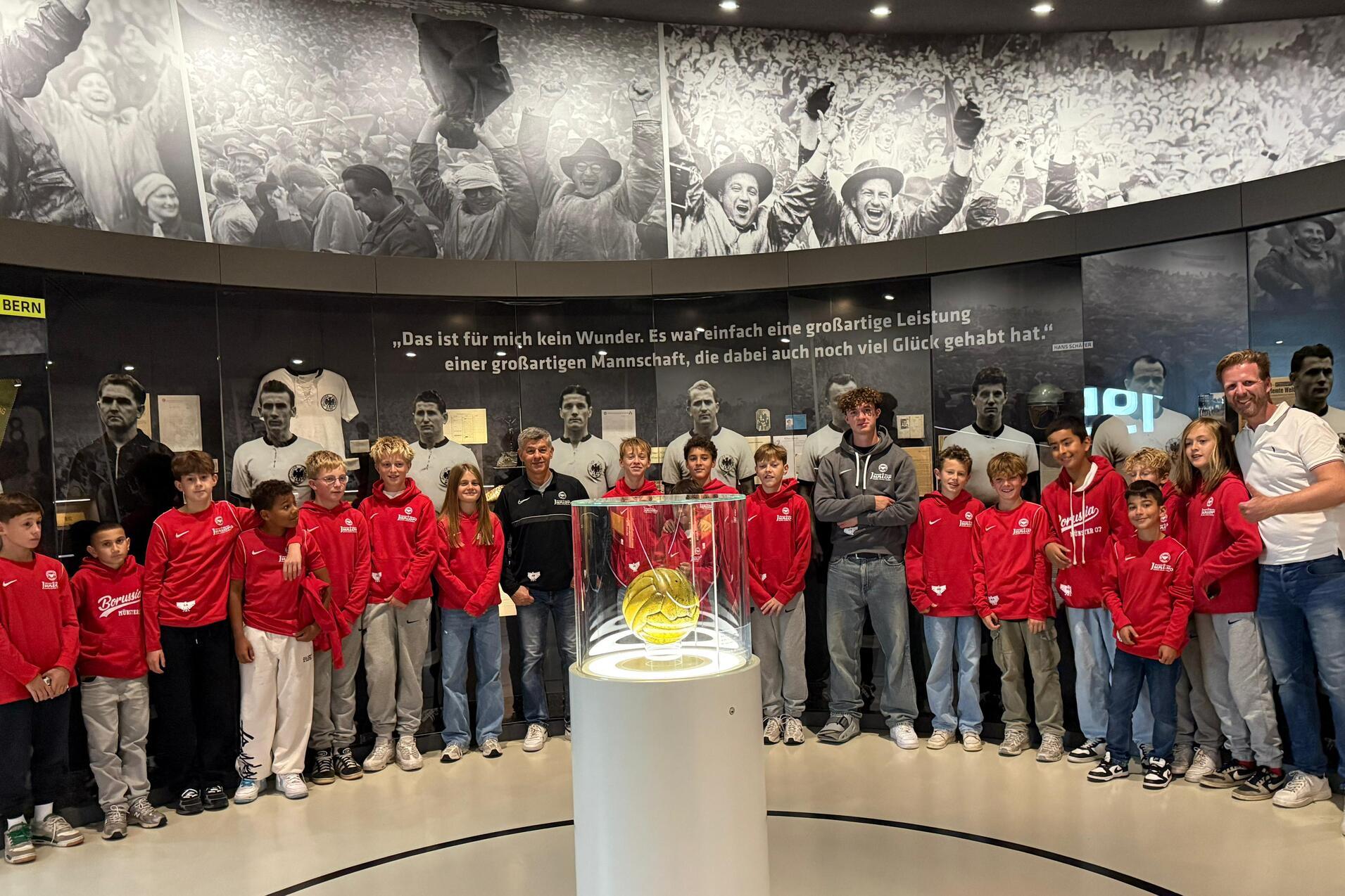 Gruppe von Kindern in roten Jacken steht um eine Glaskuppel mit einem Ball, im Hintergrund sportliche historische Bilder.