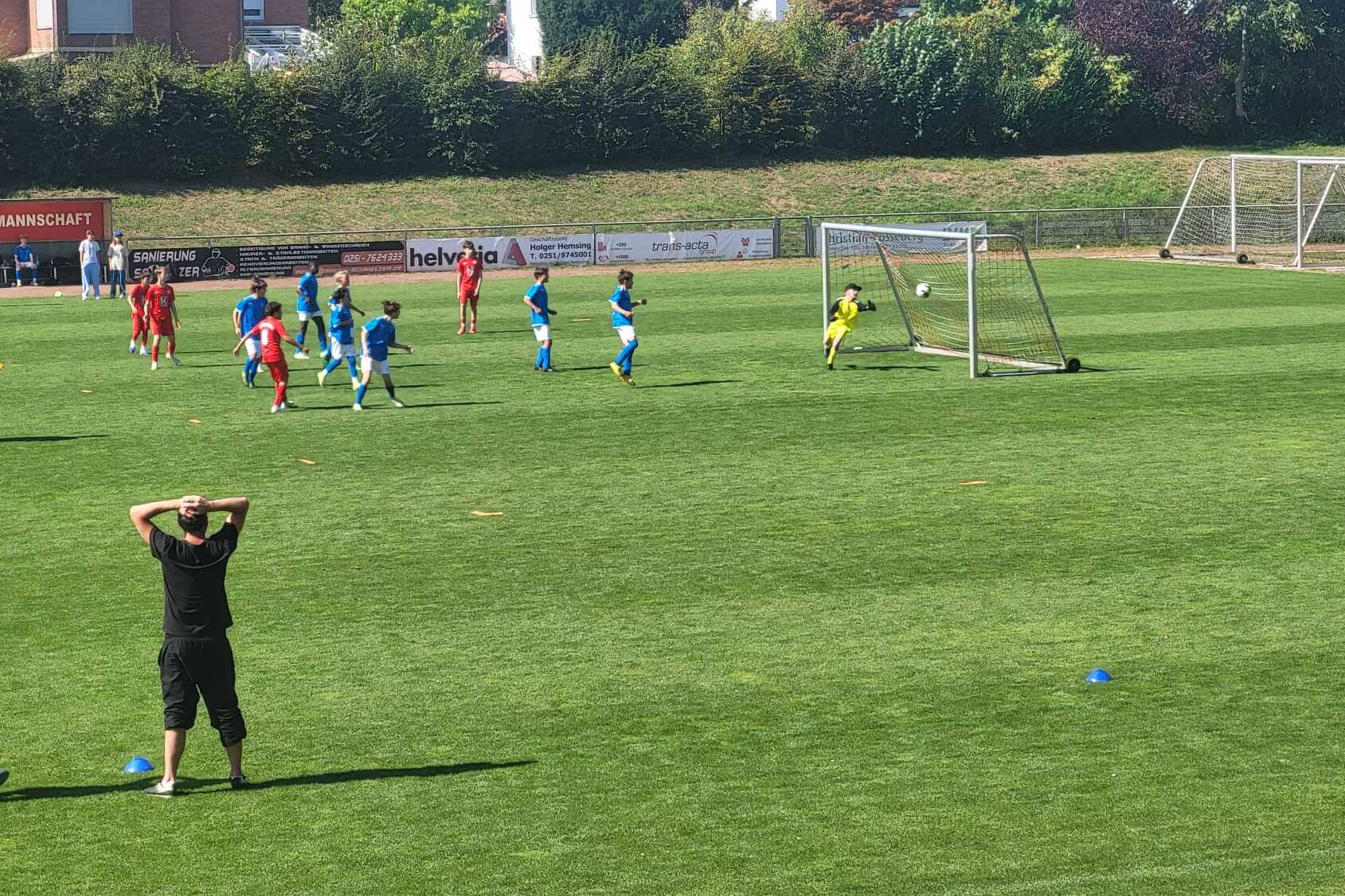Spielszene mit Kindern in blauen und roten Trikots beim Elfmeter, Zuschauer beobachten gespannt vom Spielfeldrand.