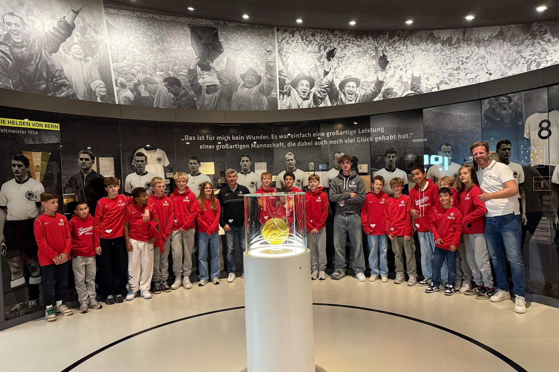 Gruppe von Kindern in roten Jacken steht um eine Glaskuppel mit einem Ball, im Hintergrund sportliche historische Bilder.