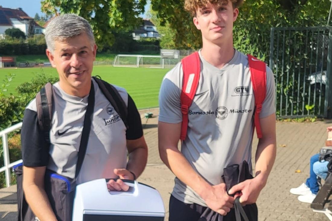 Zwei M&auml;nner in Sportkleidung stehen zusammen im Freien, der &auml;ltere mit einer K&uuml;hltasche und der j&uuml;ngere mit einem Rucksack.