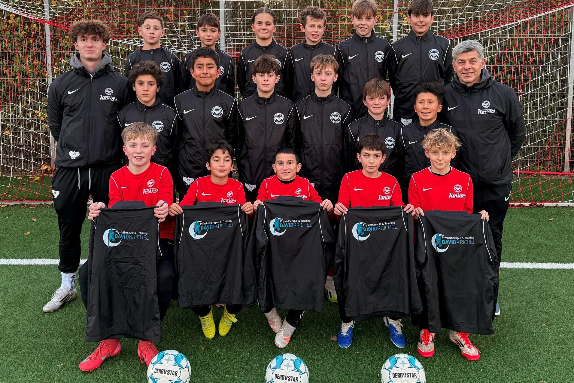 Gruppenfoto einer Jugendfu&szlig;ballmannschaft in sportlicher Kleidung, mit B&auml;llen und Trikots auf einem Fu&szlig;ballplatz.