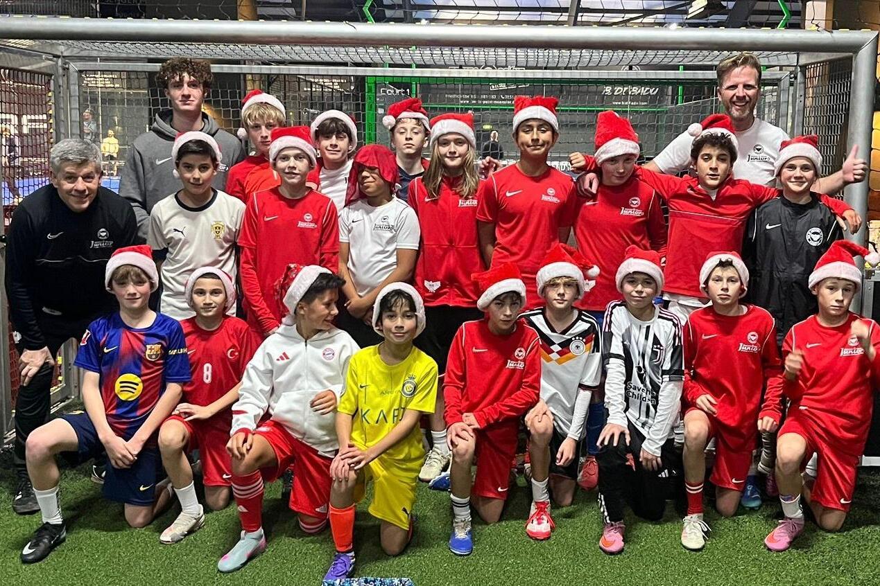 Gruppierung von 20 Kindern in Fu&szlig;balltrikots und Weihnachtsm&uuml;tzen vor einem Tor auf einem Sportplatz.