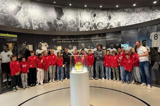 Gruppe von Kindern in roten Jacken steht um eine Glaskuppel mit einem Ball, im Hintergrund sportliche historische Bilder.