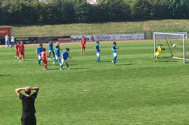 Spielszene mit Kindern in blauen und roten Trikots beim Elfmeter, Zuschauer beobachten gespannt vom Spielfeldrand.