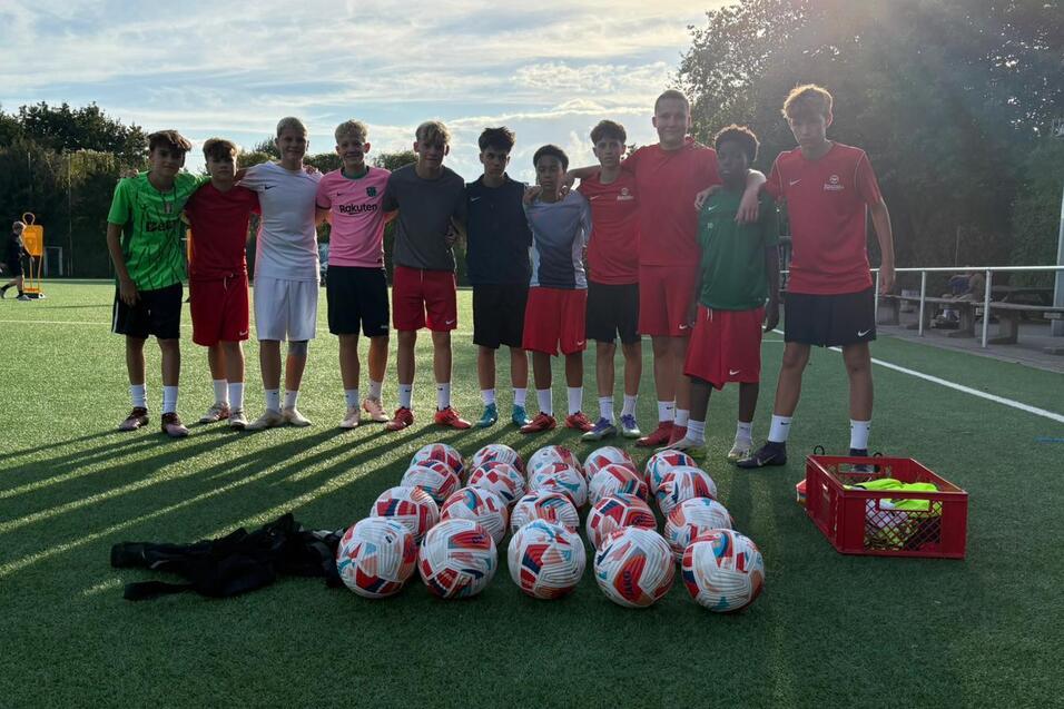 Gruppe von Fu&szlig;ballspielern in roten Trikots zusammen mit vielen Fu&szlig;ballb&auml;llen auf einem gr&uuml;nen Sportplatz.