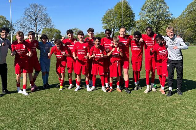 Gruppe junger Fu&szlig;ballspieler in roten Trikots posiert auf einem gr&uuml;nen Feld unter blauem Himmel.