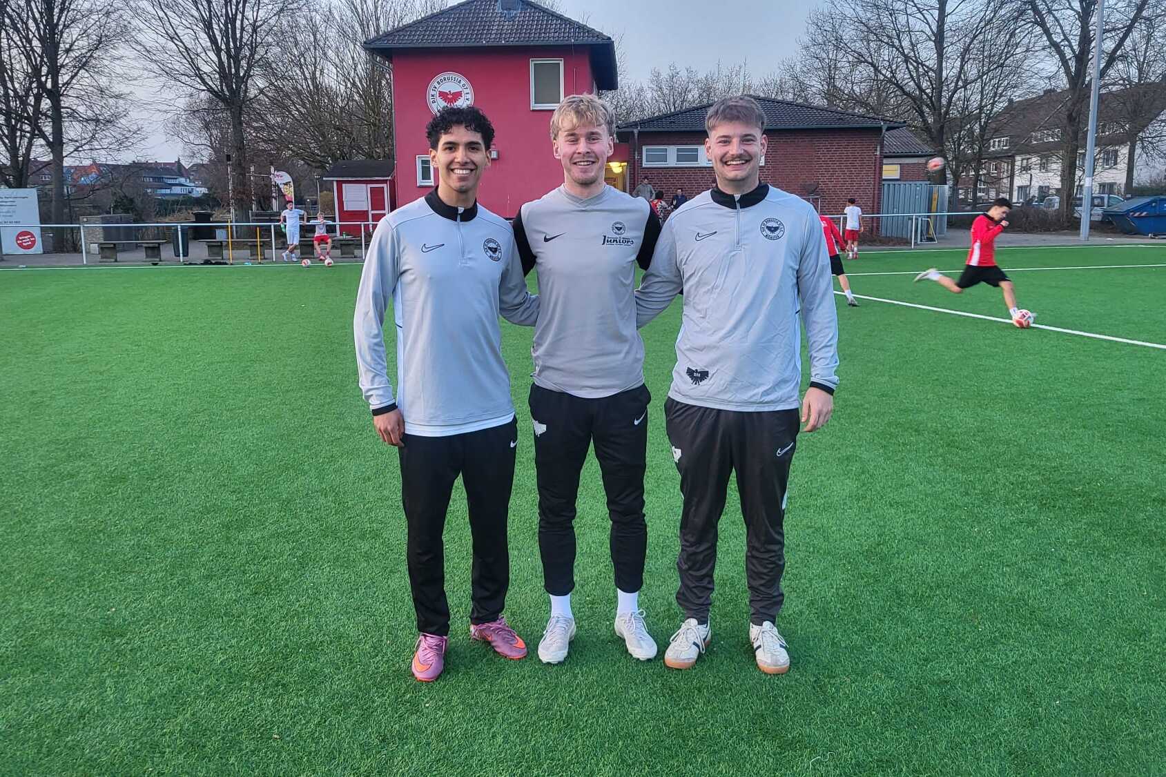 Drei Fu&szlig;ballspieler in Trainingsanz&uuml;gen stehen l&auml;chelnd auf einem Kunstrasenplatz, im Hintergrund ein Fu&szlig;ballfeld.
