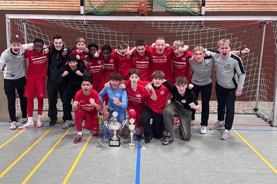 Gruppe von 15 Jugendlichen in roten Sportanz&uuml;gen posiert vor einem Fu&szlig;balltor, umgeben von Pokalen.