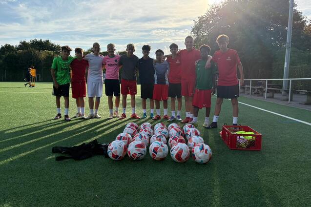 Gruppe von Fu&szlig;ballspielern in roten Trikots zusammen mit vielen Fu&szlig;ballb&auml;llen auf einem gr&uuml;nen Sportplatz.