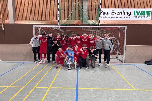 Gruppe von 15 Jugendlichen in roten Sportanz&uuml;gen posiert vor einem Fu&szlig;balltor, umgeben von Pokalen.