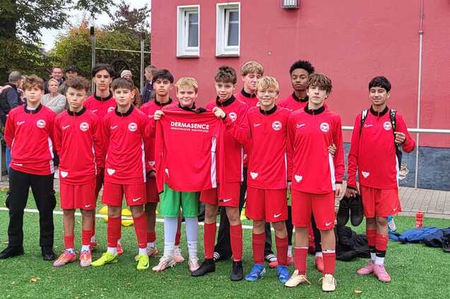 Gruppe von zw&ouml;lf Jugendlichen in roten Fu&szlig;balltrikots auf einem Kunstrasenplatz vor einem Sportgeb&auml;ude.