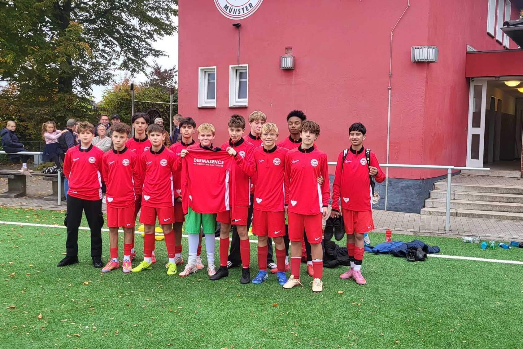Gruppe von zw&ouml;lf Jugendlichen in roten Fu&szlig;balltrikots auf einem Kunstrasenplatz vor einem Sportgeb&auml;ude.