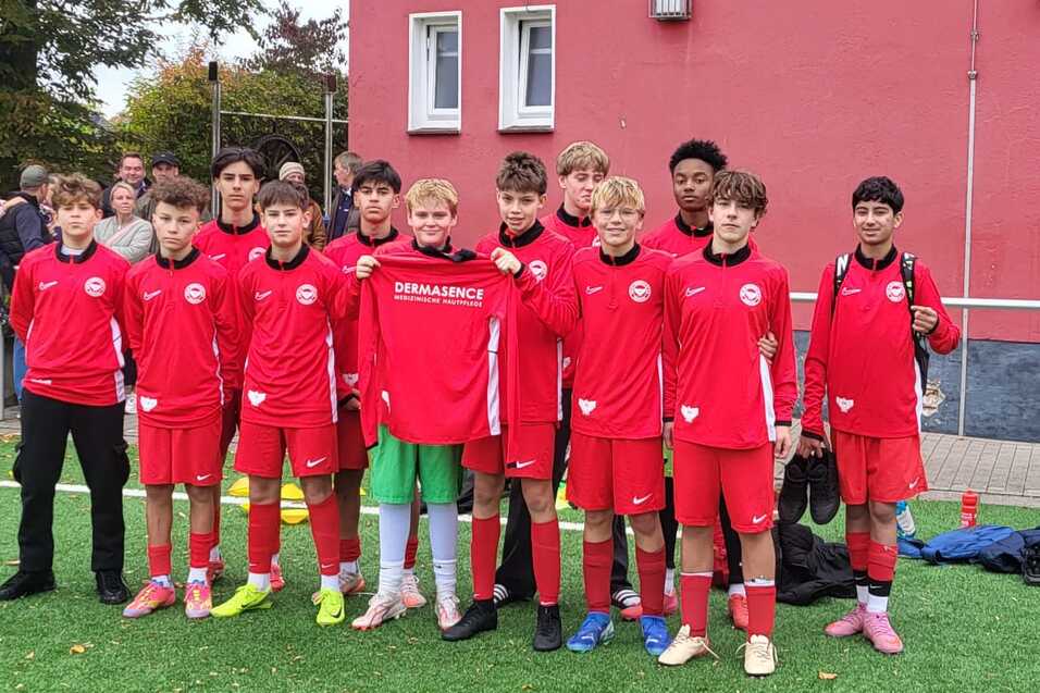 Gruppe von zw&ouml;lf Jugendlichen in roten Fu&szlig;balltrikots auf einem Kunstrasenplatz vor einem Sportgeb&auml;ude.