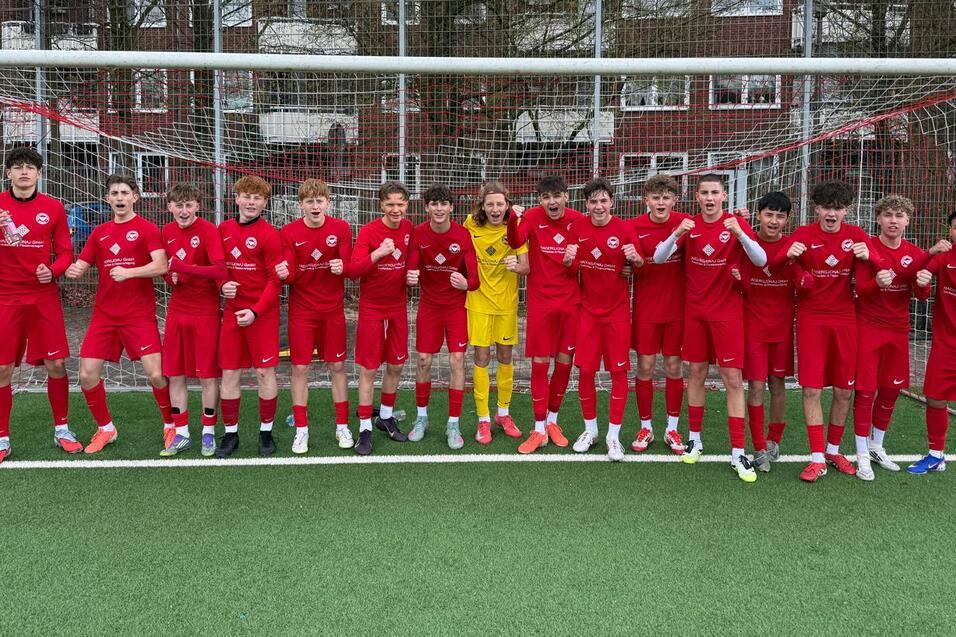 Gruppe von 17 Fu&szlig;ballspielern in roten Trikots, posierend vor einem Tor auf einem Kunstrasenplatz.
