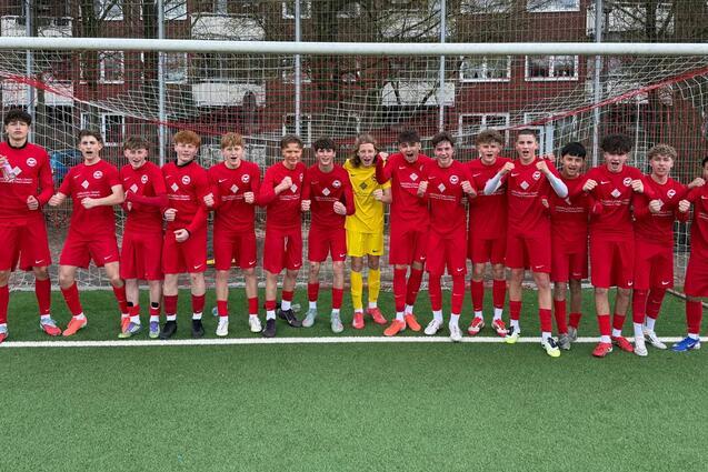 Gruppe von 17 Fu&szlig;ballspielern in roten Trikots, posierend vor einem Tor auf einem Kunstrasenplatz.
