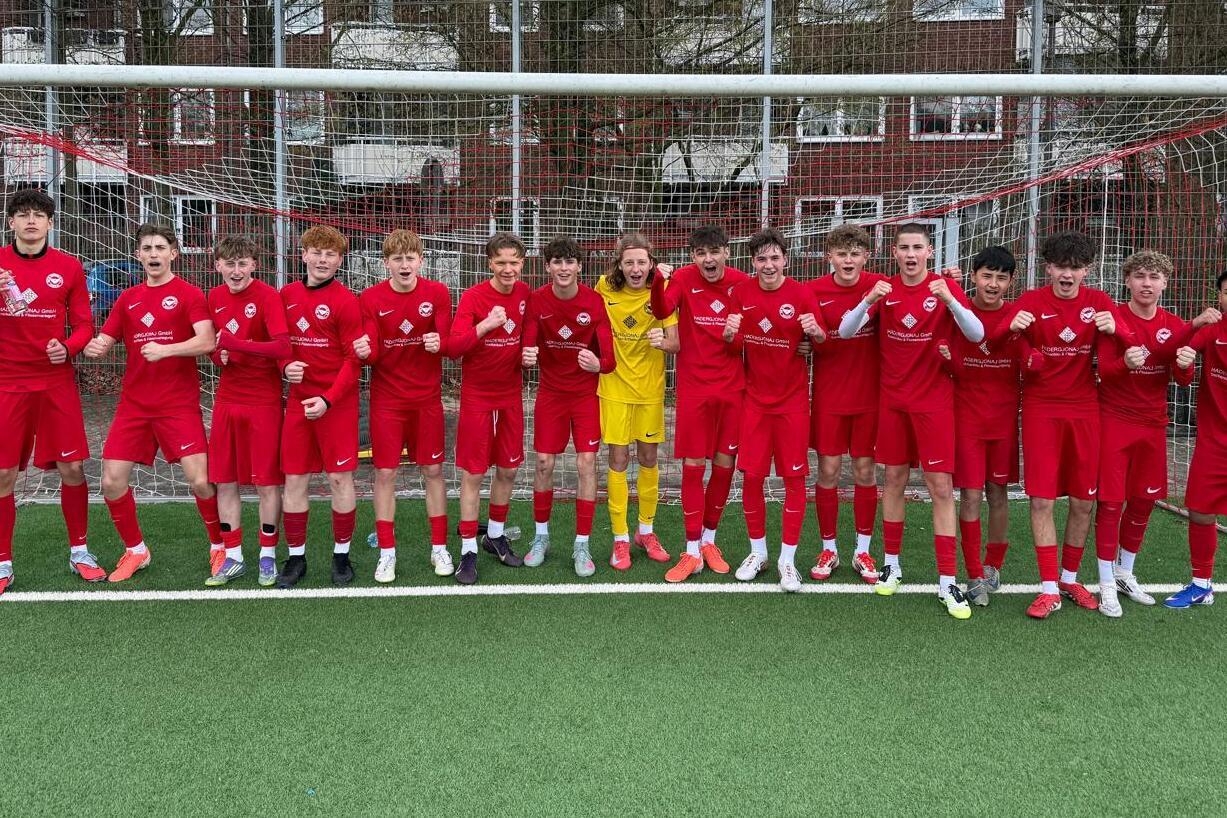 Gruppe von 17 Fußballspielern in roten Trikots, posierend vor einem Tor auf einem Kunstrasenplatz.