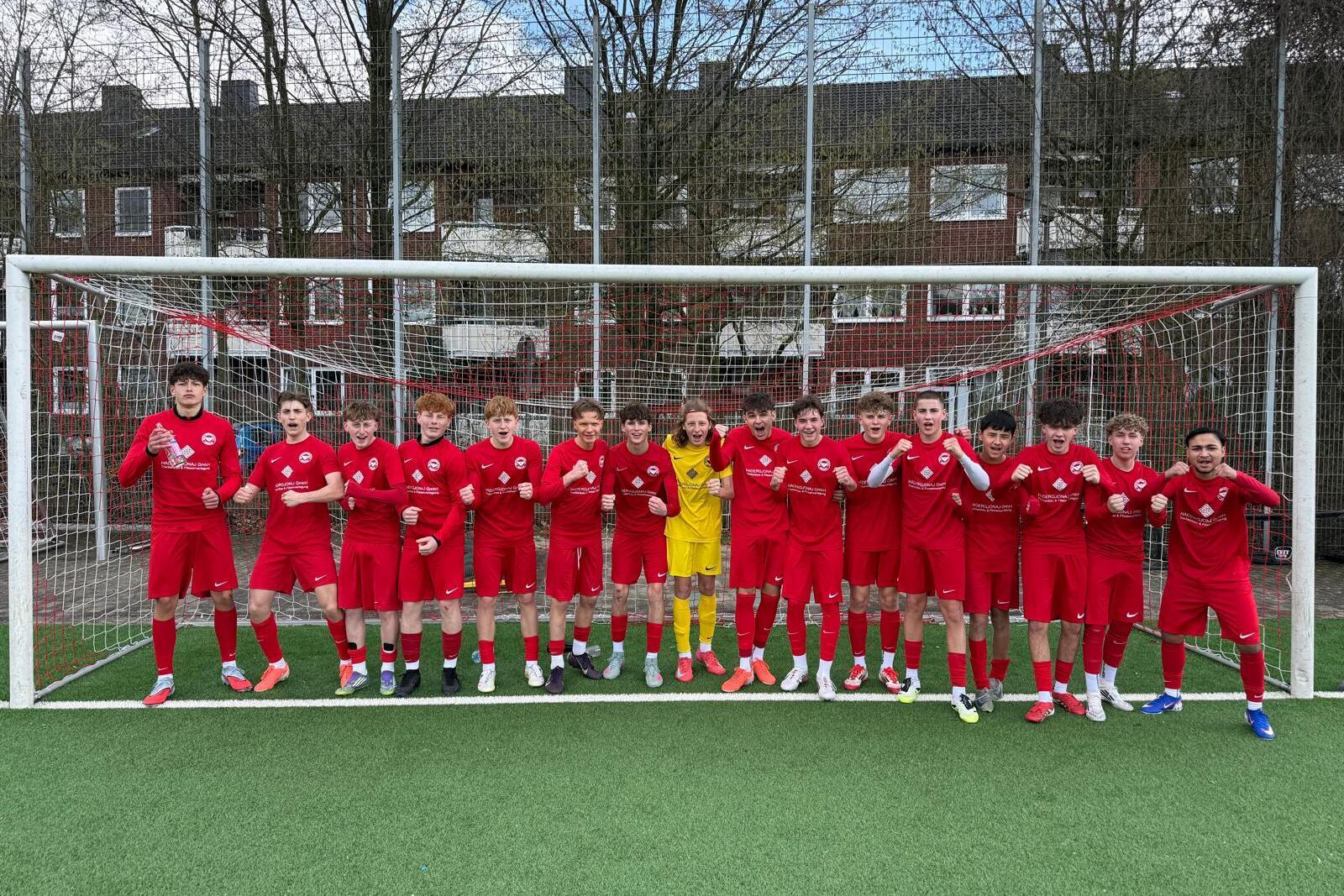 Gruppe von 17 Fu&szlig;ballspielern in roten Trikots, posierend vor einem Tor auf einem Kunstrasenplatz.