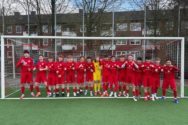 Gruppe von 17 Fu&szlig;ballspielern in roten Trikots, posierend vor einem Tor auf einem Kunstrasenplatz.