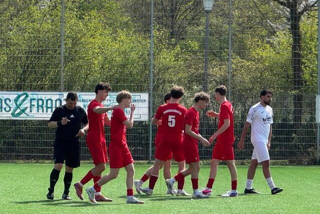 Sechs Fu&szlig;ballspieler in roten Trikots stehen auf einem Kunstrasenplatz, w&auml;hrend ein Schiedsrichter in Schwarz daneben steht.