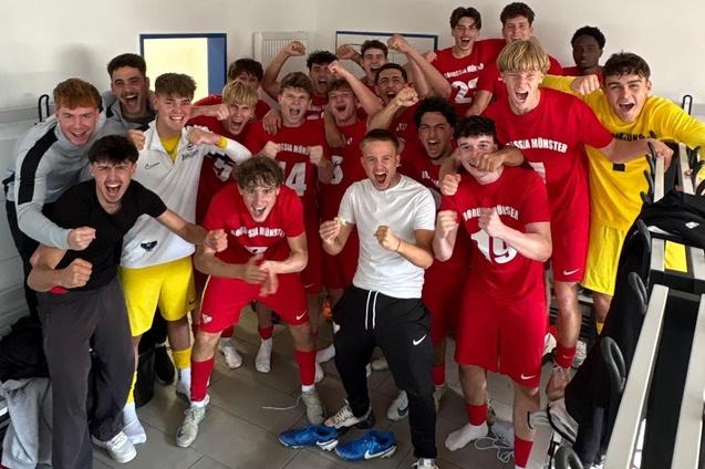 Gruppe von jubelnden Fu&szlig;ballspielern in roten Trikots vor einer Umkleidekabine, die ihren Sieg feiern.
