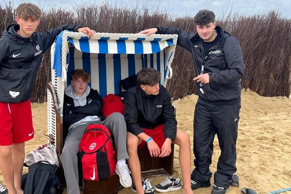 Vier Jugendliche stehen am Strand, einer sitzt in einem Strandkorb, alle tragen sportliche Kleidung und Flip-Flops.