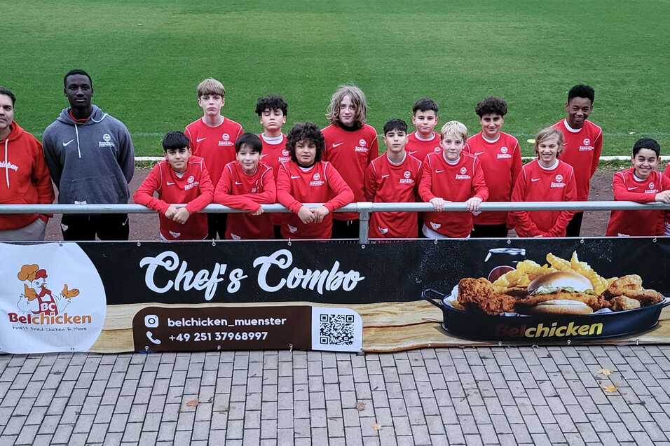 Gruppe von Fu&szlig;ballspielern in roten Trikots posiert mit Trainern vor einer Werbebande auf einem Sportplatz.