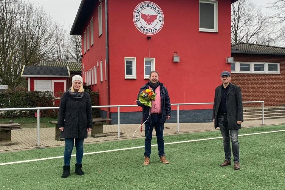 Drei Personen stehen auf einem Fu&szlig;ballplatz vor einem roten Vereinsgeb&auml;ude, einer davon h&auml;lt einen Blumenstrau&szlig;.
