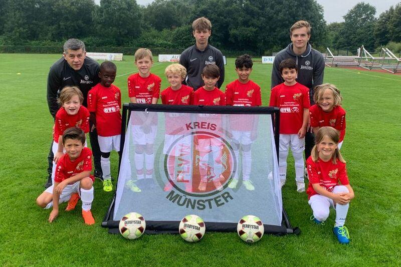 Gruppe von Fu&szlig;ballspielern in roten Trikots posiert vor einem Banner mit dem Schriftzug &bdquo;KREIS M&Uuml;NSTER&ldquo; auf einem Rasenplatz.