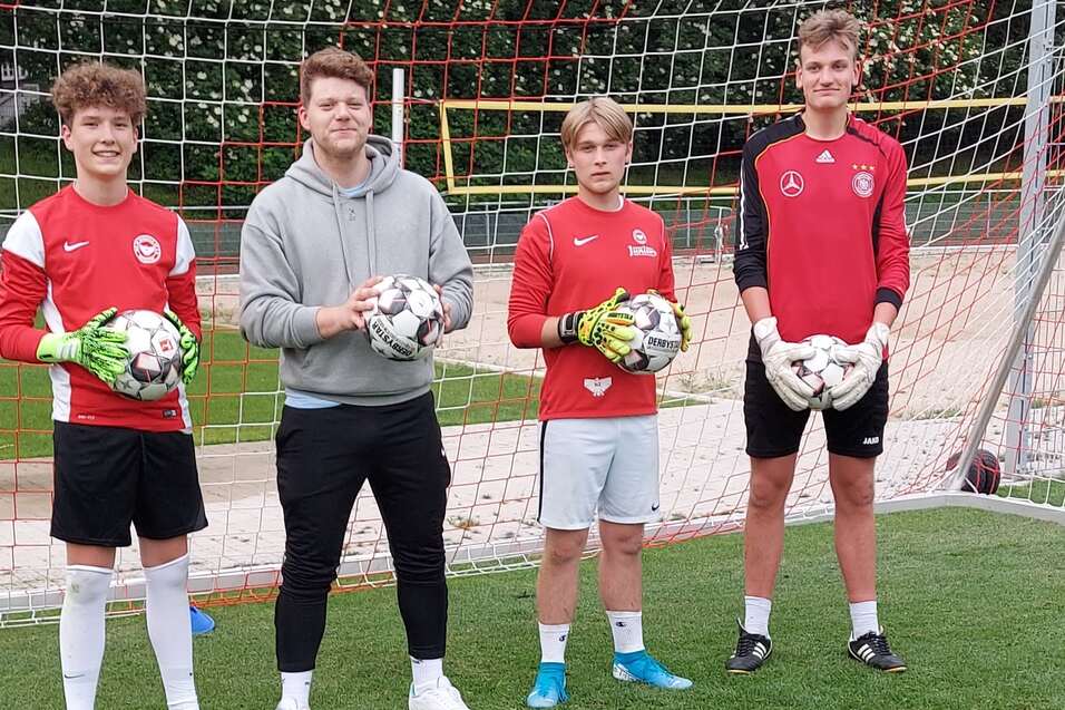 Drei Fu&szlig;ballspieler in roten Trikots stehen mit B&auml;llen vor einem Fu&szlig;balltor auf einem Rasenplatz.