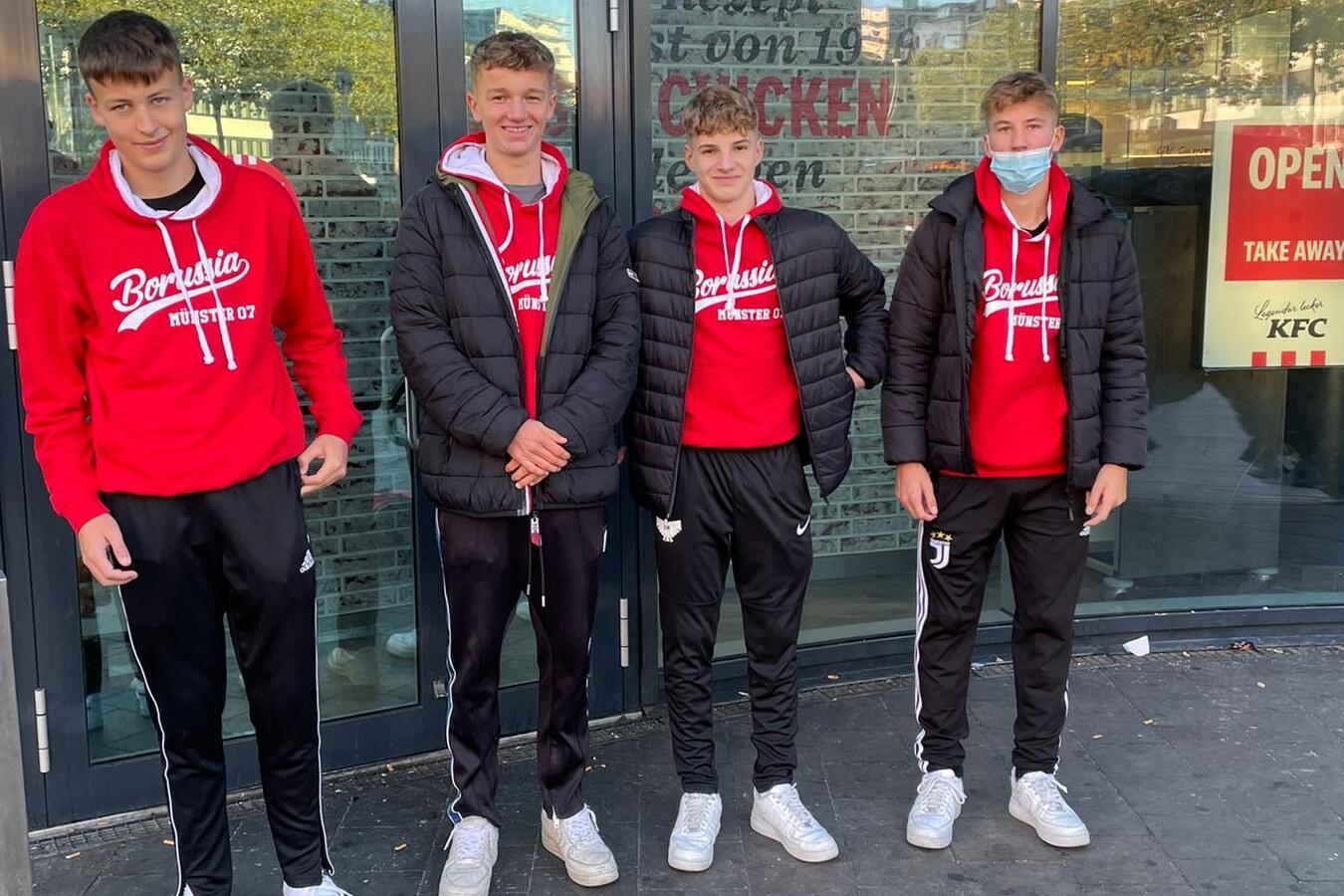 Vier Jugendliche in roten Sweatshirts und Sporthosen stehen vor einem Restaurant mit gro&szlig;en Fenstern.