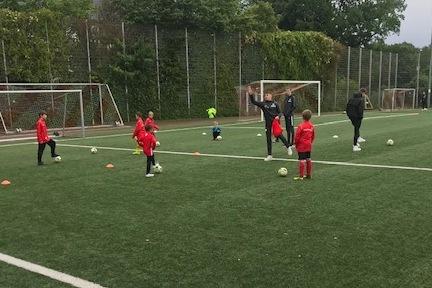 Gruppe von Kindern in roten Trainingsanz&uuml;gen &uuml;bt mit Fu&szlig;b&auml;llen auf einem Fu&szlig;ballfeld mit Kunstrasen.