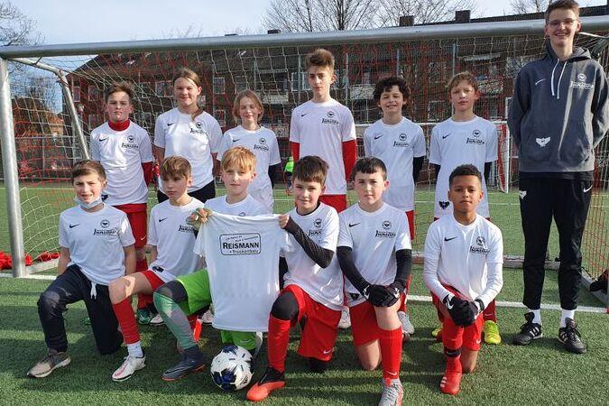 Gruppe von zw&ouml;lf Jugendlichen in wei&szlig;en Shirts mit roten Hosen, posierend vor einem Fu&szlig;balltor auf einem Platz.