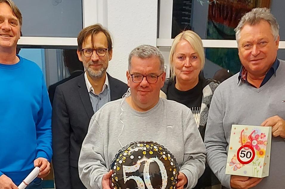 F&uuml;nf Personen stehen zusammen, eine Person h&auml;lt einen schwarzen Luftballon mit der Zahl 50, im Hintergrund sind Geschenke.