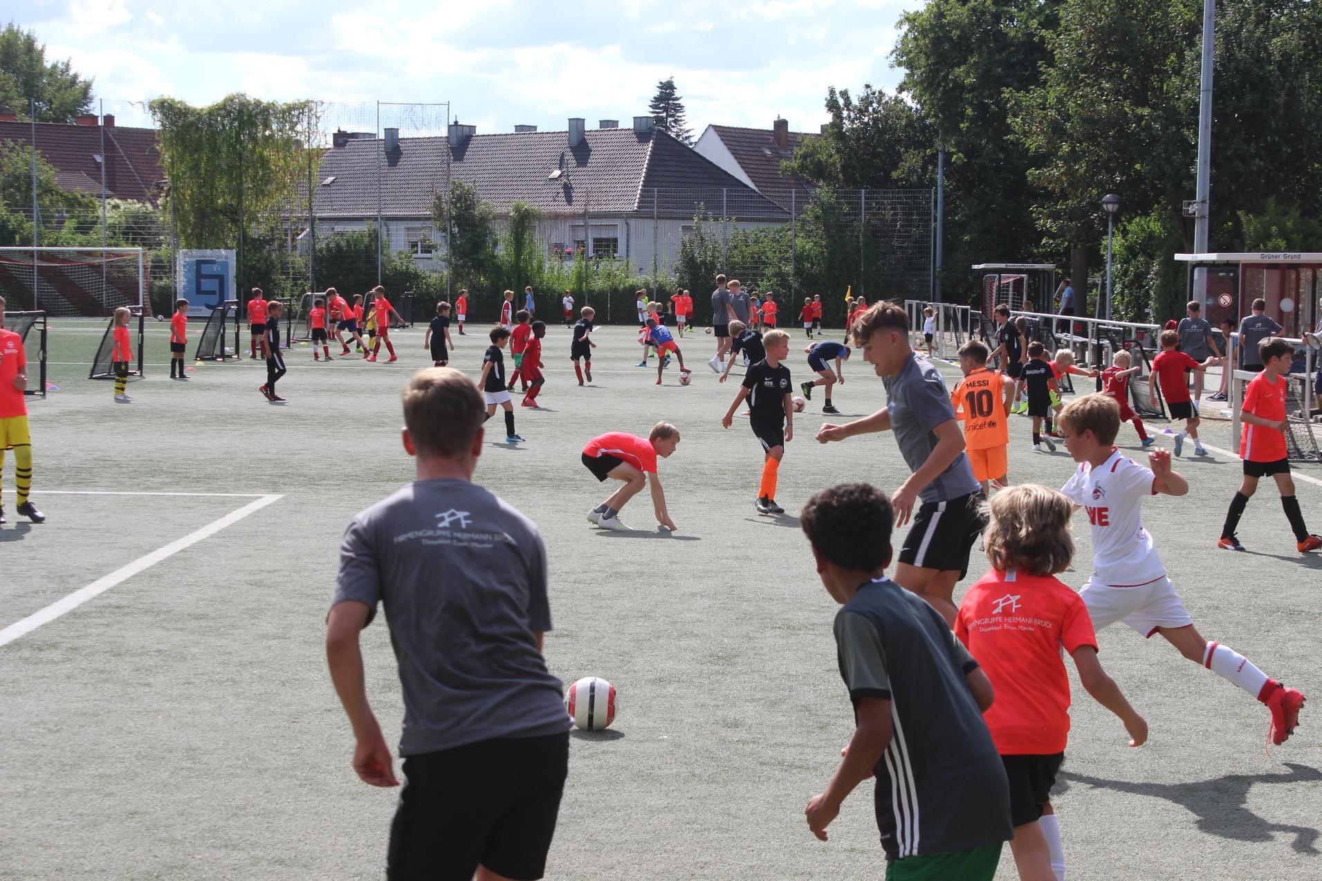 Gruppentraining von Kindern auf einem Fu&szlig;ballplatz, viele Spieler in bunten Shirts und Shorts, &uuml;ber verschiedene Aktivit&auml;ten verteilt.