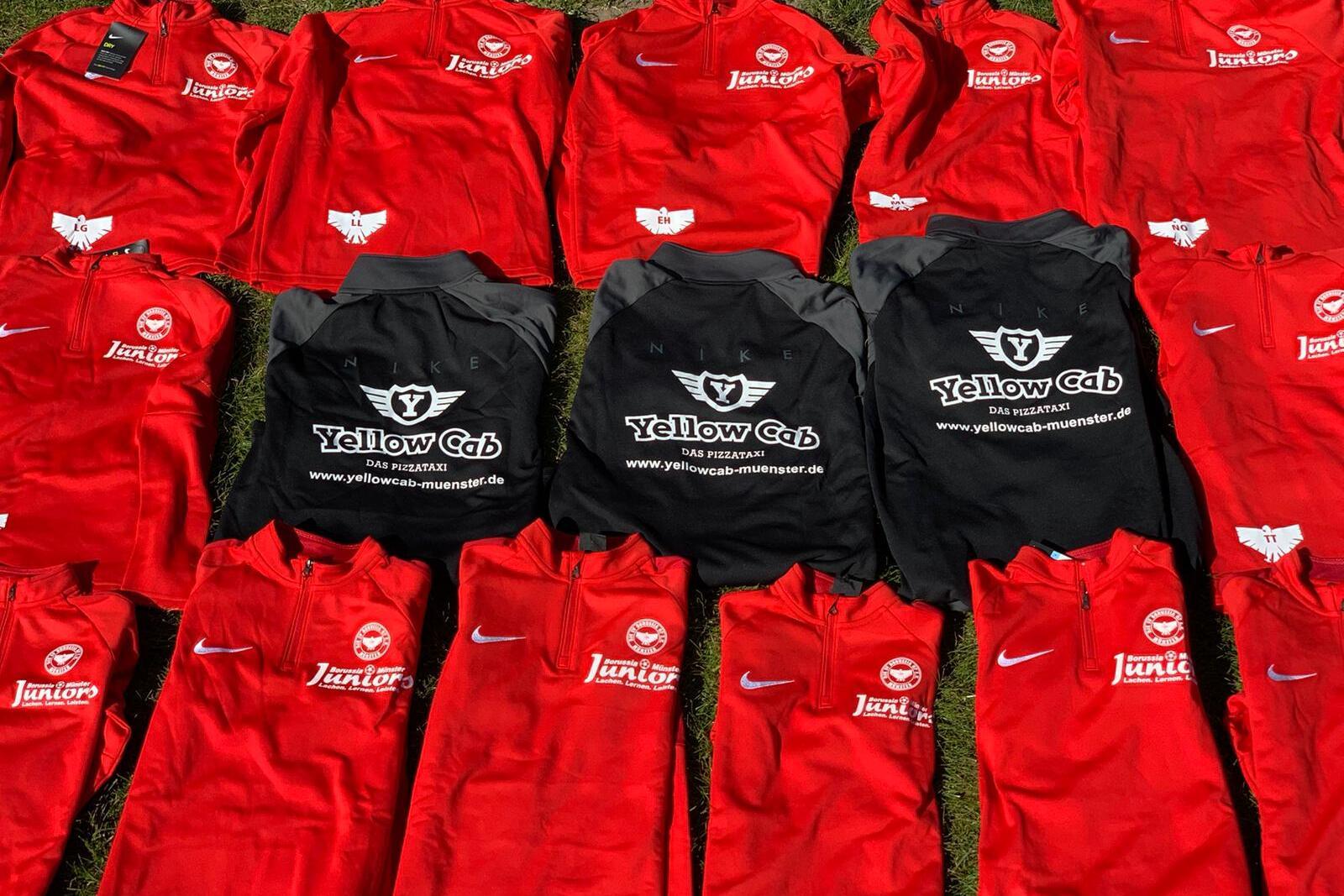Eine Anordnung roter Trikots und schwarzer T-Shirts auf gr&uuml;nem Gras, einige mit Logos von Sponsoren.