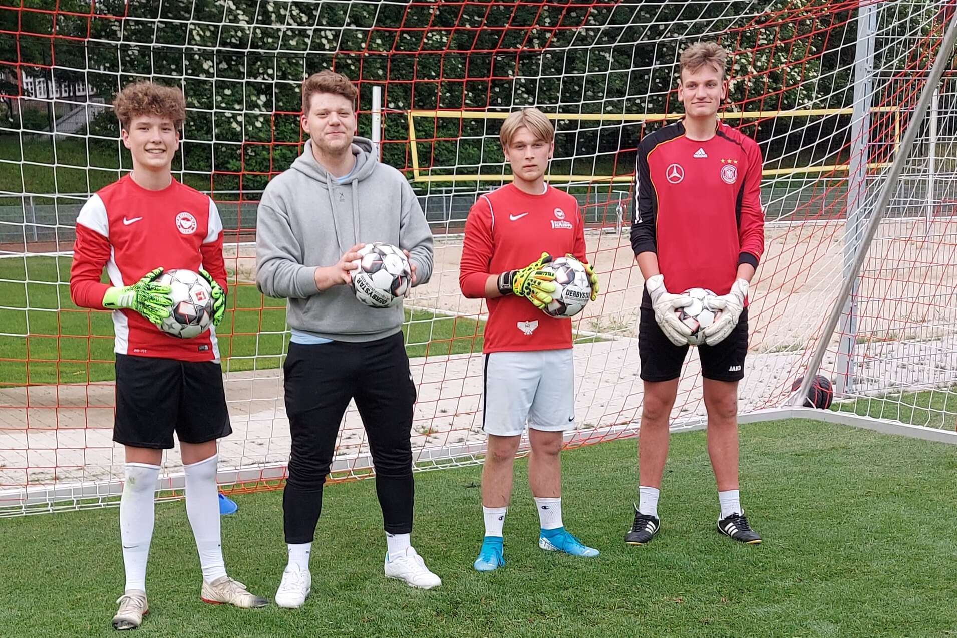 Drei Fu&szlig;ballspieler in roten Trikots stehen mit B&auml;llen vor einem Fu&szlig;balltor auf einem Rasenplatz.