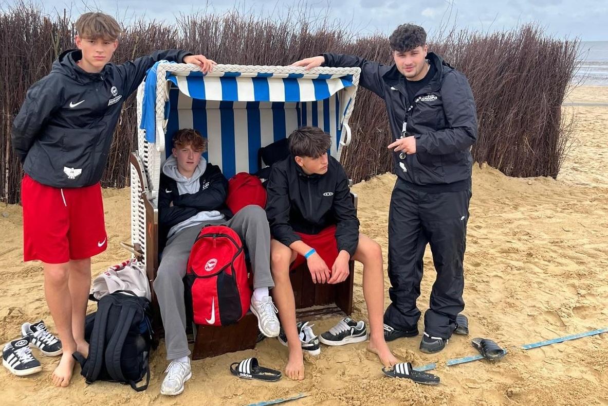 Vier Jugendliche stehen am Strand, einer sitzt in einem Strandkorb, alle tragen sportliche Kleidung und Flip-Flops.