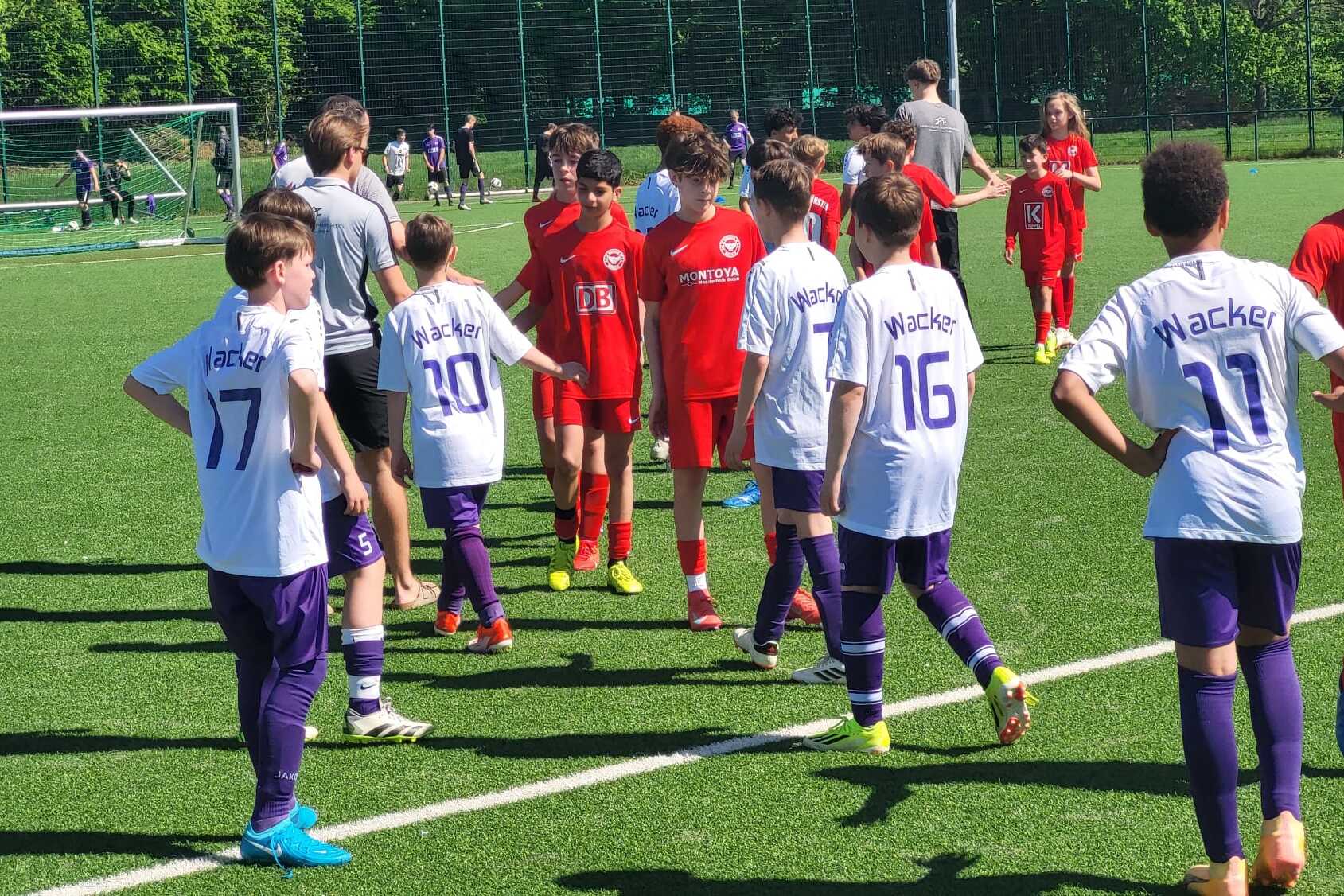 Zwei Gruppen von Jugendlichen in Fu&szlig;balltrikots stehen auf einem Kunstrasenplatz und sprechen miteinander.