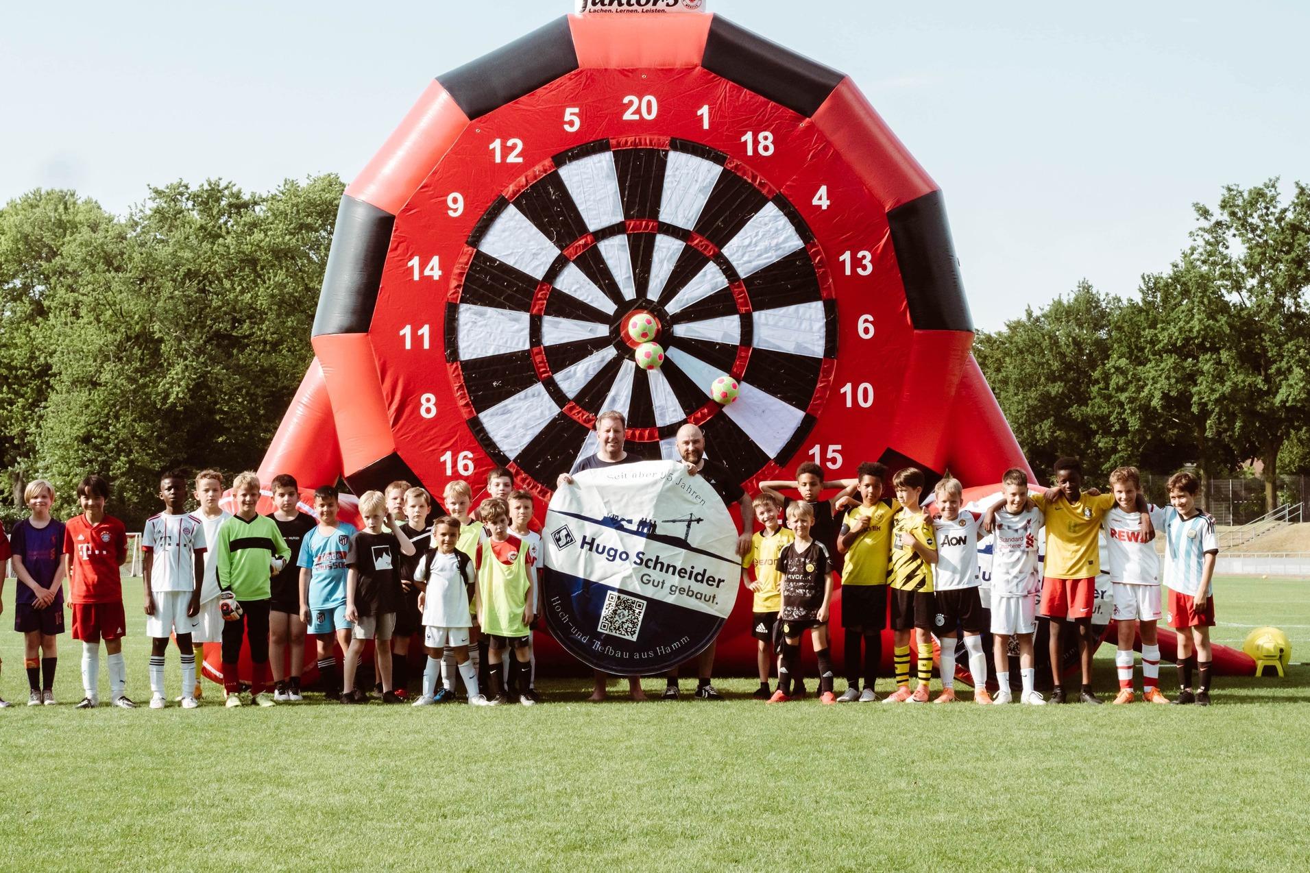 Gruppierung von Kindern im Fu&szlig;balloutfit vor einer gro&szlig;en, bunten Dartscheibe auf einem Sportplatz.