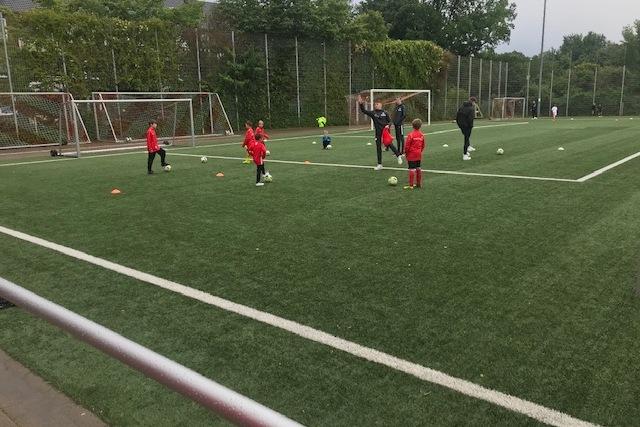 Gruppe von Kindern in roten Trainingsanz&uuml;gen &uuml;bt mit Fu&szlig;b&auml;llen auf einem Fu&szlig;ballfeld mit Kunstrasen.
